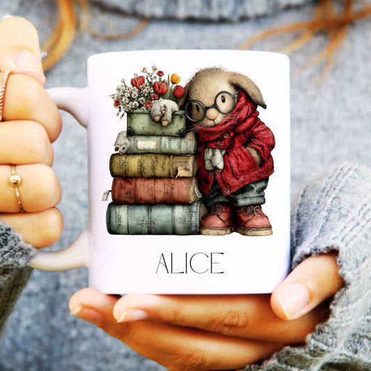 Mug with a design of a bookish rabbit character holding books and flowers, with 'ALICE' text, held by hands.