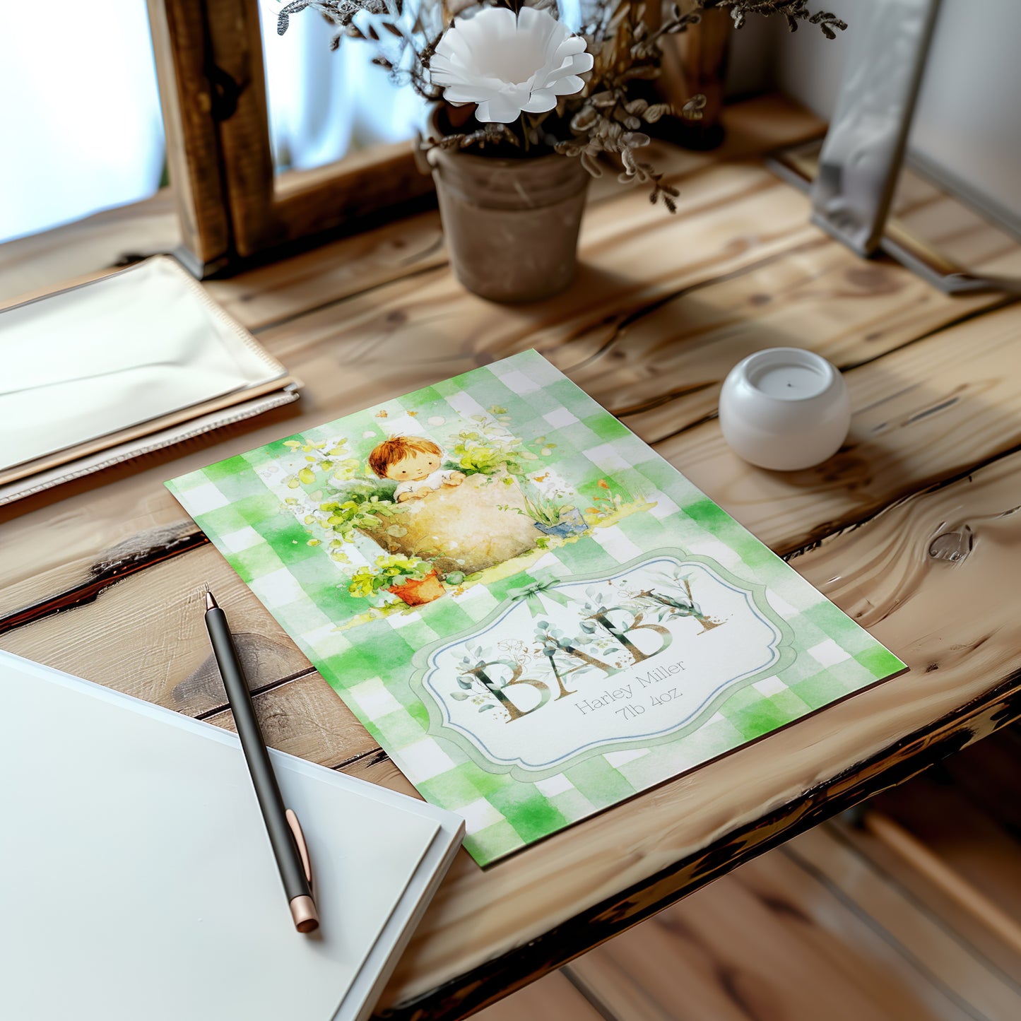 A baby card with a green and white gingham pattern and a baby illustration, styled on a rustic wooden table next to a notebook, pen, and small candle.