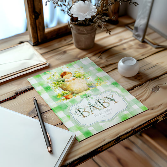 A baby card with a green and white gingham pattern and a baby illustration, styled on a rustic wooden table next to a notebook, pen, and small candle.