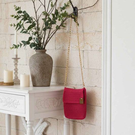 Pink woven handbag hanging on a hook next to a vase with greenery on a white table.