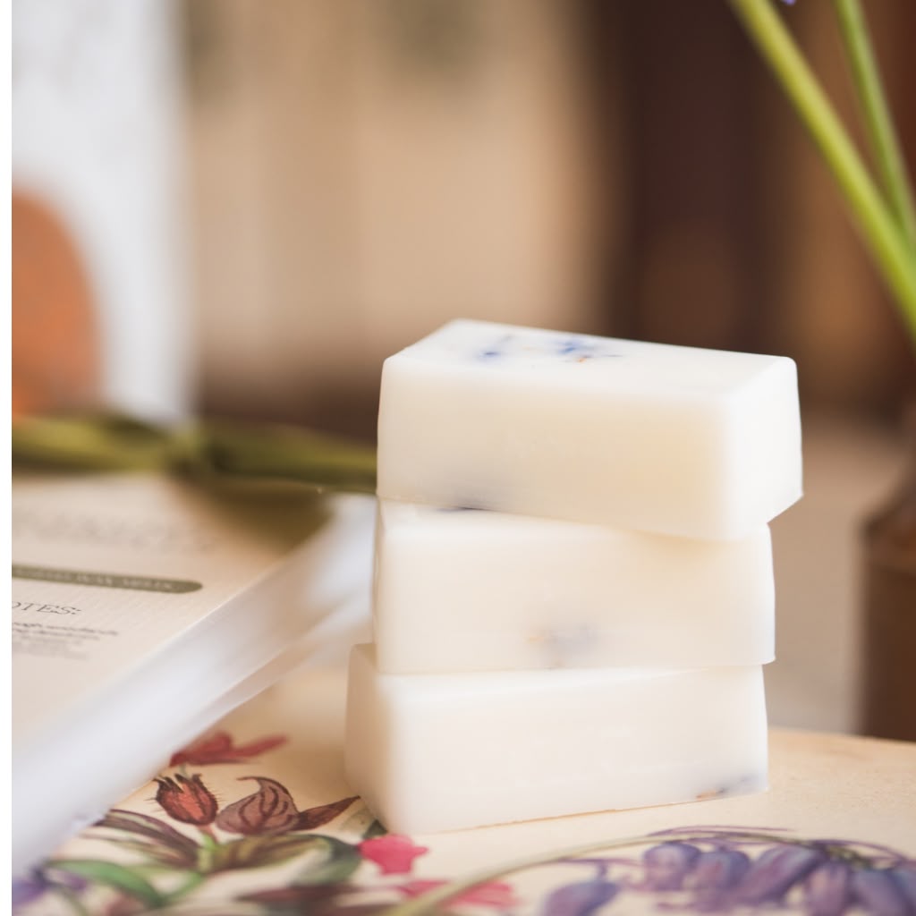 Three stacked rectangular bars of creamy off-white wax melts with small blue floral inclusions, displayed on a floral surface with a blurred background.