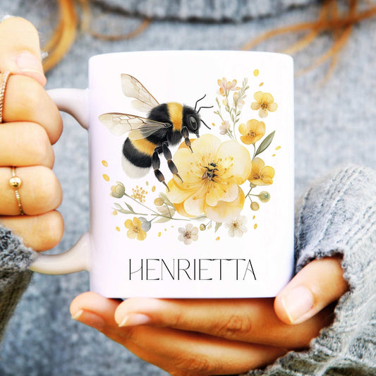 Person holding a mug with a bee and floral design, personalised with 'HENRIETTA'.