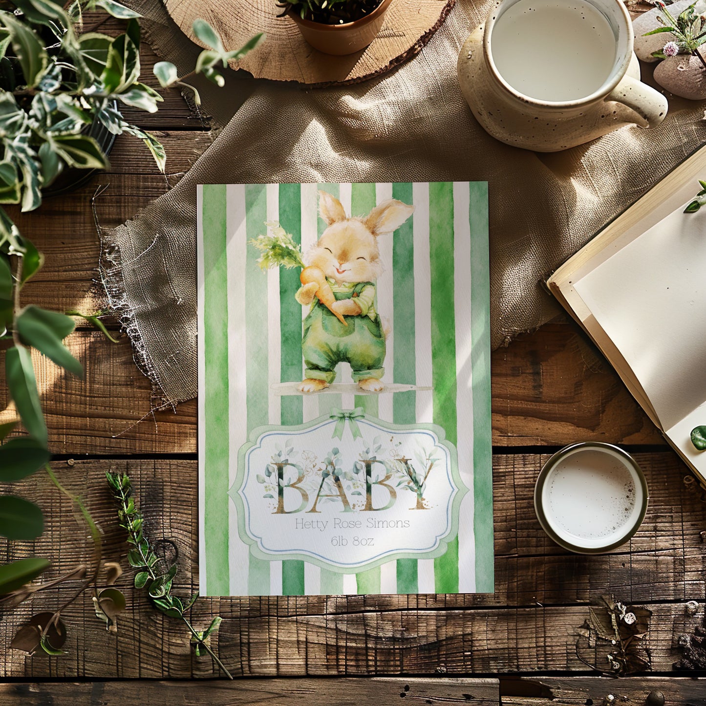 A baby card laid flat on a rustic wooden table, surrounded by decorative props including green plants, a mug of milk, an open book, and a small candle. The bag has green and white vertical stripes, a watercolour bunny illustration, and the text 'BABY'