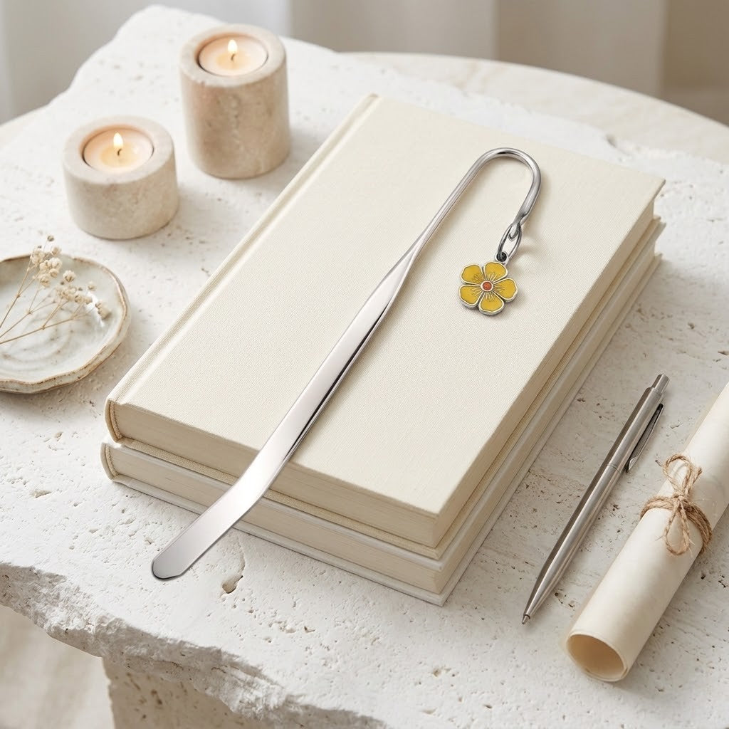 A silver metal bookmark shaped like a small yellow flower with pink and blue centre details, resting on a stack of cream-coloured notebooks on a textured white stone surface, with candles, a pen, and a rolled paper scroll nearby.