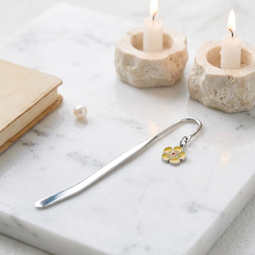 A silver metal bookmark shaped like a flower with yellow and white enamel petals and a small pink centre, resting on a white marble surface with candles, a book, and a pearl nearby.