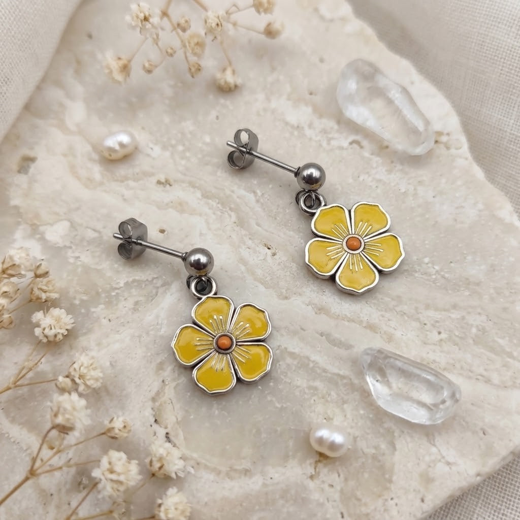 A pair of yellow flower dangle earrings with silver-toned posts and backings, displayed on a textured beige stone slab with dried flowers and clear crystals.