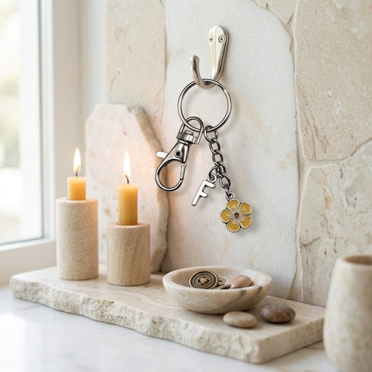 A silver metal keyring with a key and a small yellow flower charm hanging on a beige stone wall hook. The scene is styled with two lit candles, a small stone dish, and stones on a stone tray near a window.