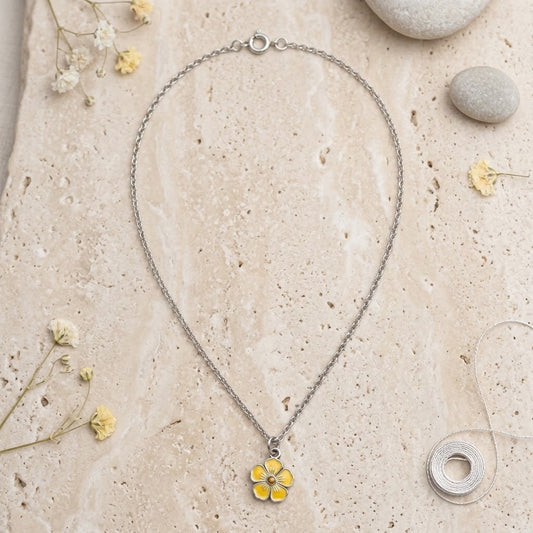 A delicate silver chain necklace with a small yellow flower pendant, styled on a textured beige stone background with dried flowers and small stones.
