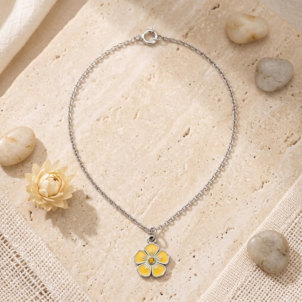 A delicate silver chain necklace with a small yellow flower charm, styled on a textured beige stone slab with small decorative stones and fabric.