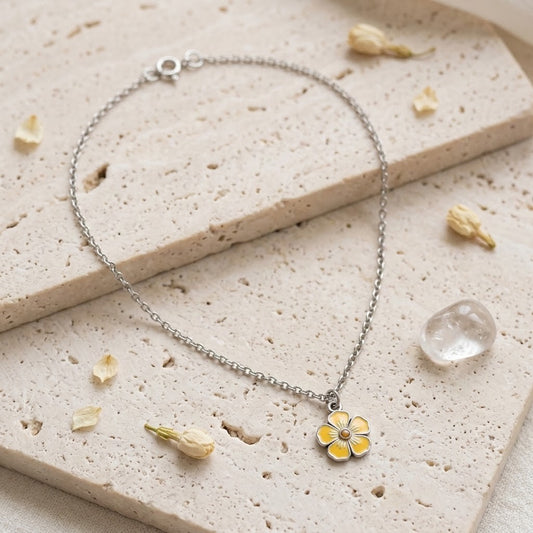 A delicate silver chain bracelet with a small yellow flower charm, styled on a textured beige stone slab with dried flower petals and a clear crystal.