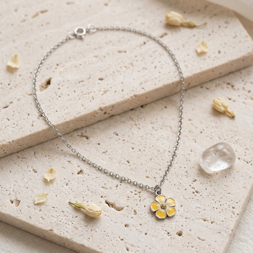 A delicate silver chain bracelet with a small yellow flower charm, styled on a textured beige stone slab with dried flower petals and a clear crystal.