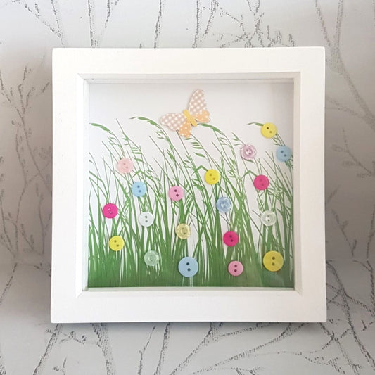 A framed button art picture featuring a butterfly and colorful meadow flowers in a white frame.