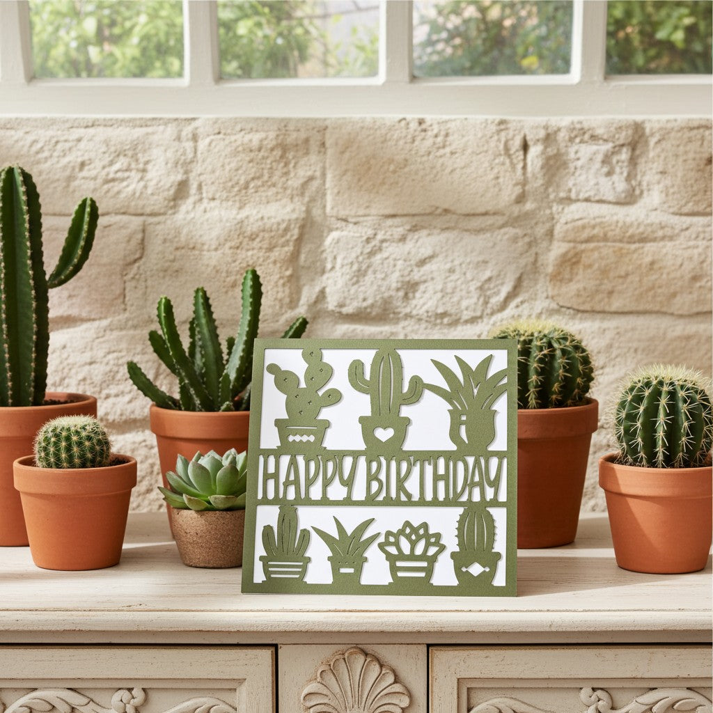 A square white card with olive green cactus and succulent silhouettes and the words “HAPPY BIRTHDAY,” displayed on a light wood cabinet in a styled room with potted cacti and a stone wall background.