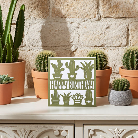 A square white card with a sage green cactus and plant pattern and the words “HAPPY BIRTHDAY” in the centre, displayed on a white cabinet in front of a stone wall with potted cacti.