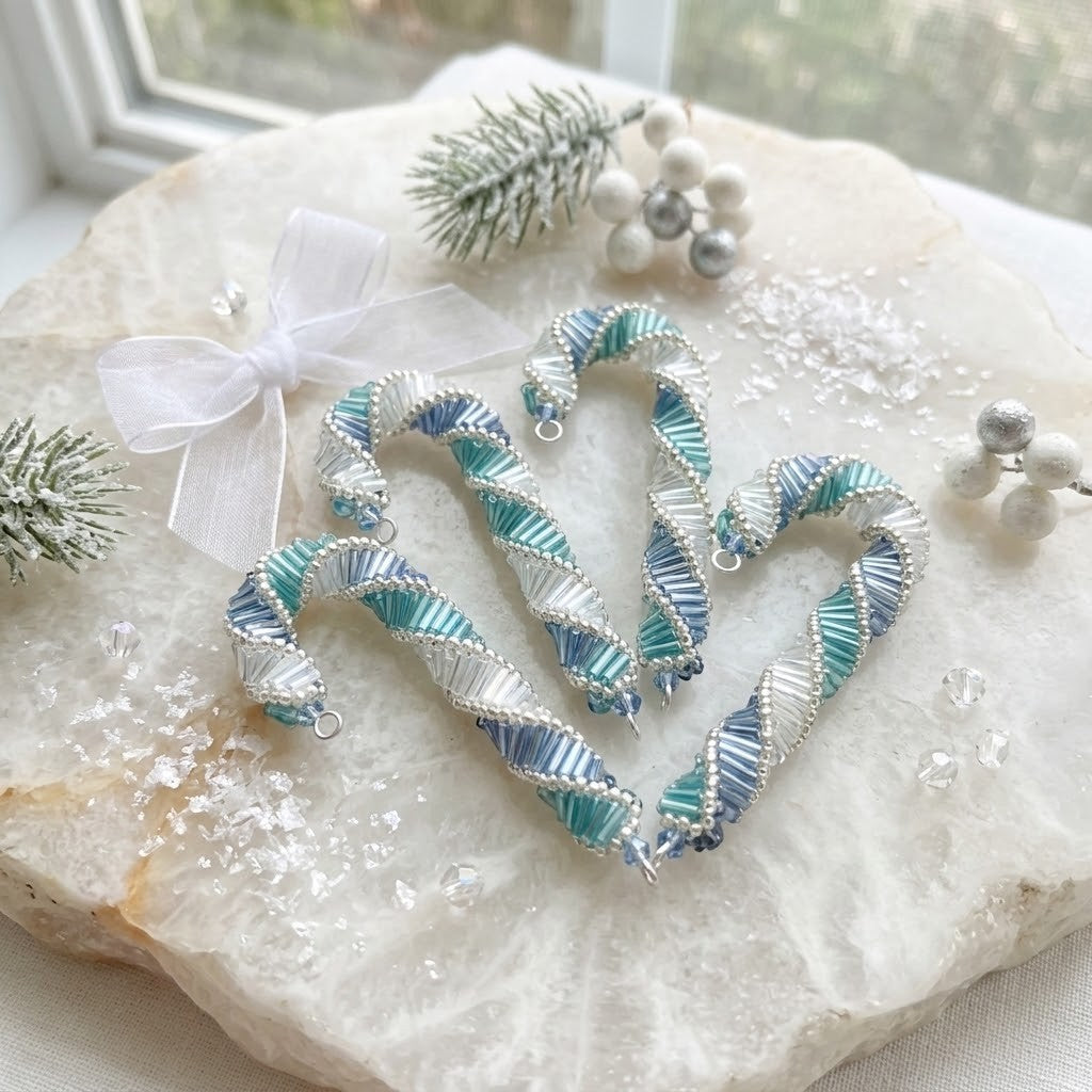 Four handmade beaded candy canes in a blue, teal, and silver spiral design, arranged on a white crystal slab with a white ribbon bow, miniature pine branches, and silver beads.