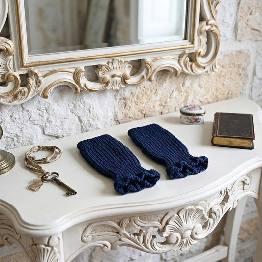 A pair of dark navy blue ribbed knit fingerless gloves warmers with ruffled cuffs, displayed on an ornate white console table in a styled room setting with a mirror, keys, and a book.