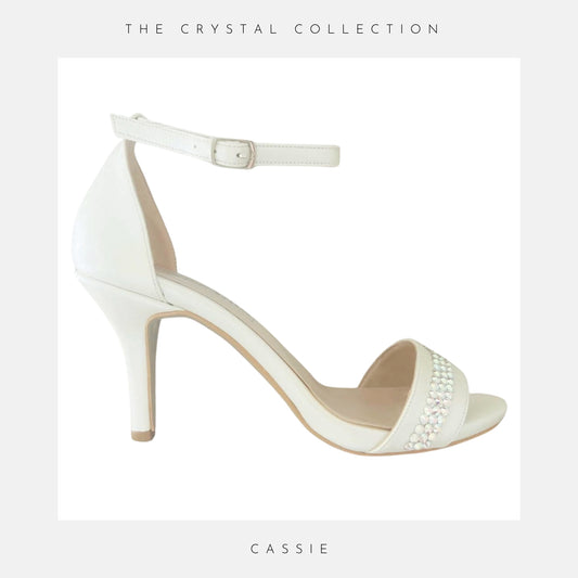 Ivory sandal with ankle strap and pearl embellishments on a light grey background, labelled 'Cassie' from 'The Crystal Collection'.