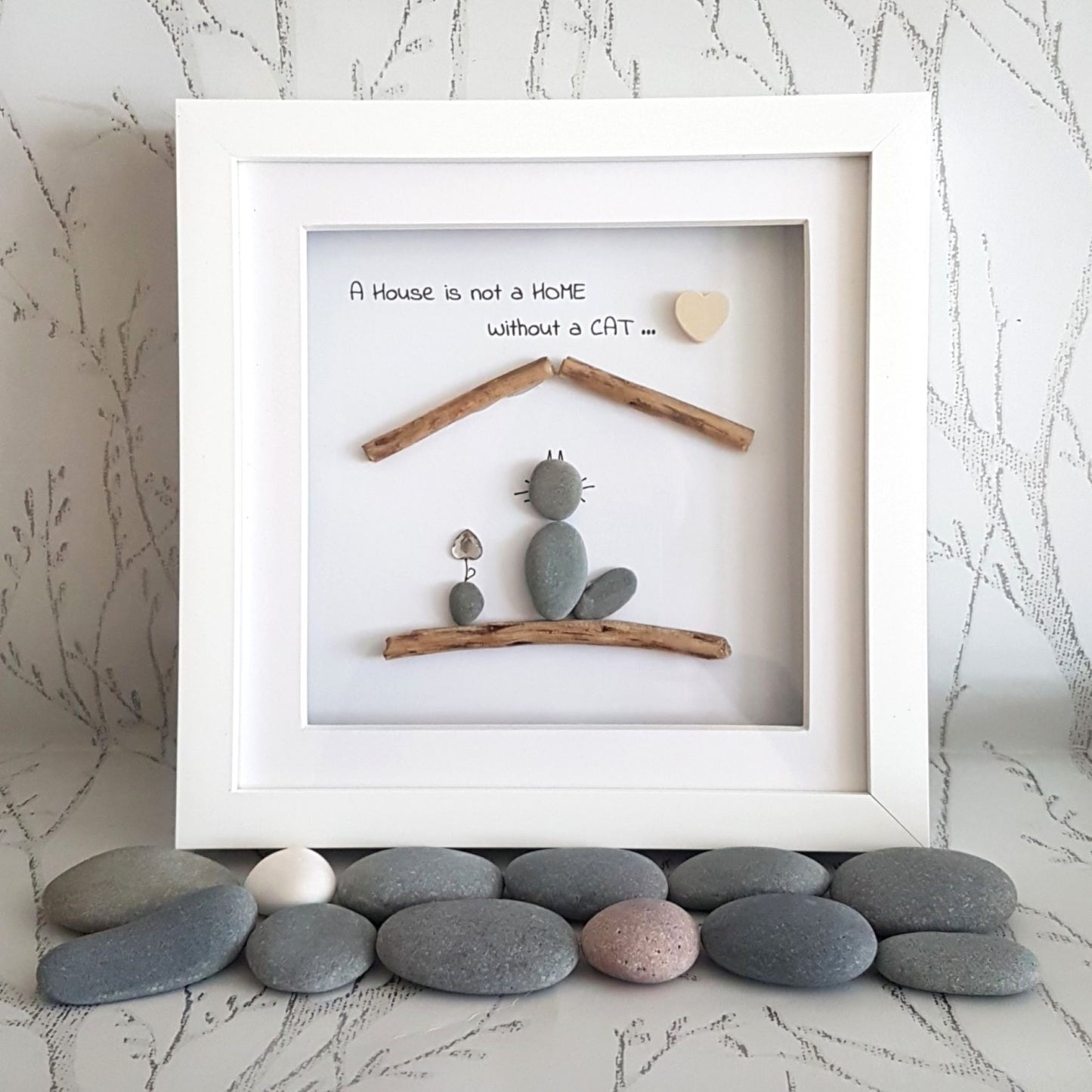 A decorative pebble art picture frame with a cat figure, a house, and a heart-shaped cut-out, displayed in a white frame, with a quote 'A house is not a home without a cat.'.
