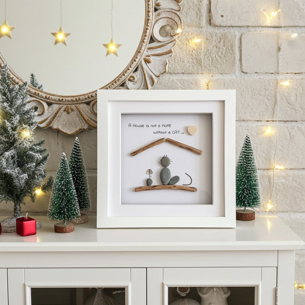 A white square shadow box framed piece of wall art sits on a white cabinet, decorated with small green Christmas trees, a red candle, and warm string lights against a brick wall.