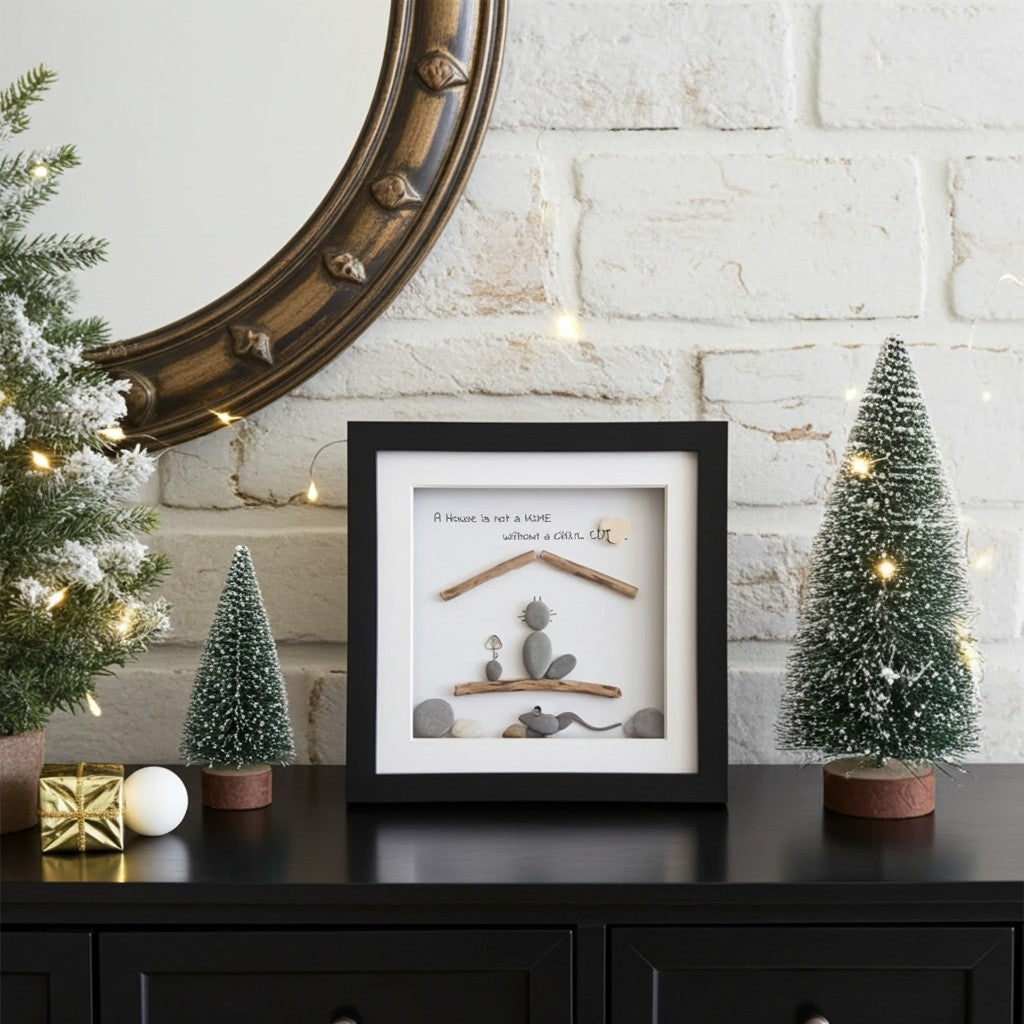 A framed shadow box art piece with a black frame and white mat, featuring a small dog silhouette, wooden branches, stones, and the text 'A House is not a Home without a pet.' It is styled on a black cabinet against a white brick wall, flanked by miniature Christmas trees and warm string lights.