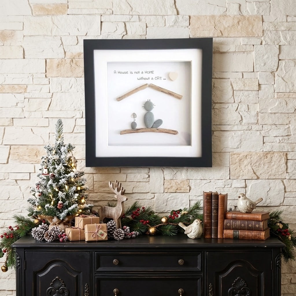Framed wall art with a black frame and white mat, featuring a small cat and the quote 'A house is not a home without a cat', displayed above a dark wooden console table with holiday decor against a light stone wall.