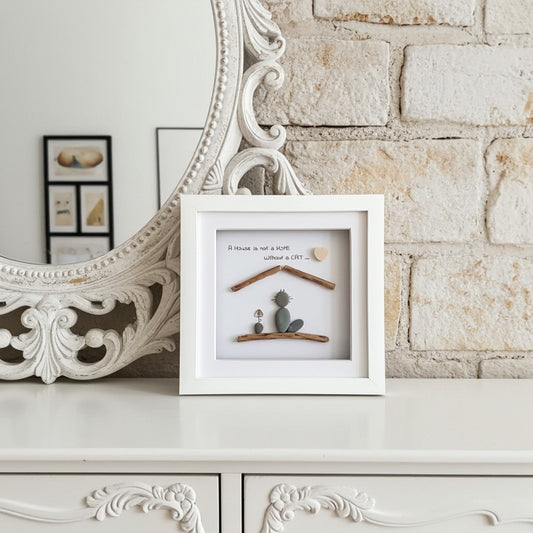 A white square shadow box frame containing an owner and a cat made from pebbles with a lovely quote