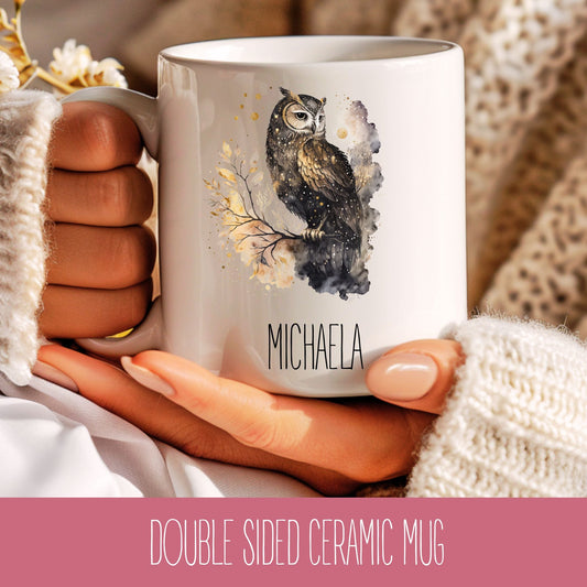 A person holding a white ceramic coffee mug with a printed owl design and the name 'MICHAELA' on it, wearing a beige sweater.