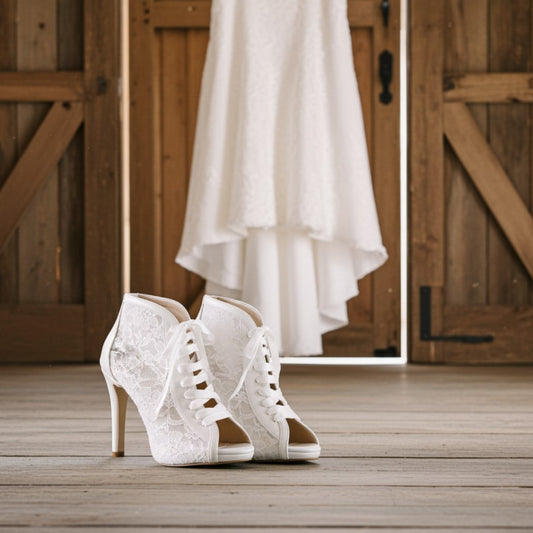 Ivory lace wedding boots on a wooden floor with a white dress hanging in the background.