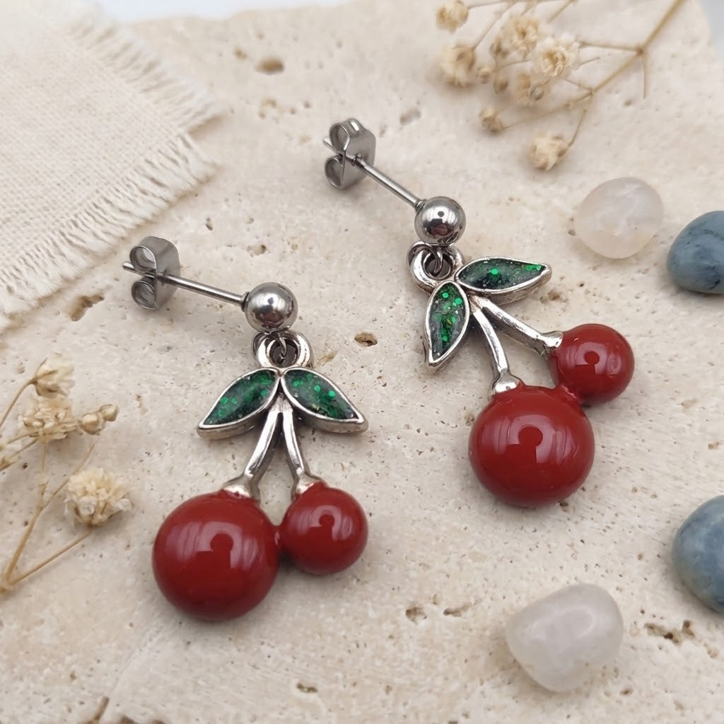A pair of cherry-shaped stud earrings with polished silver-toned metal posts, green leaf inlays, and two red round bead cherries, displayed on a textured beige stone surface with small dried flowers and stones.