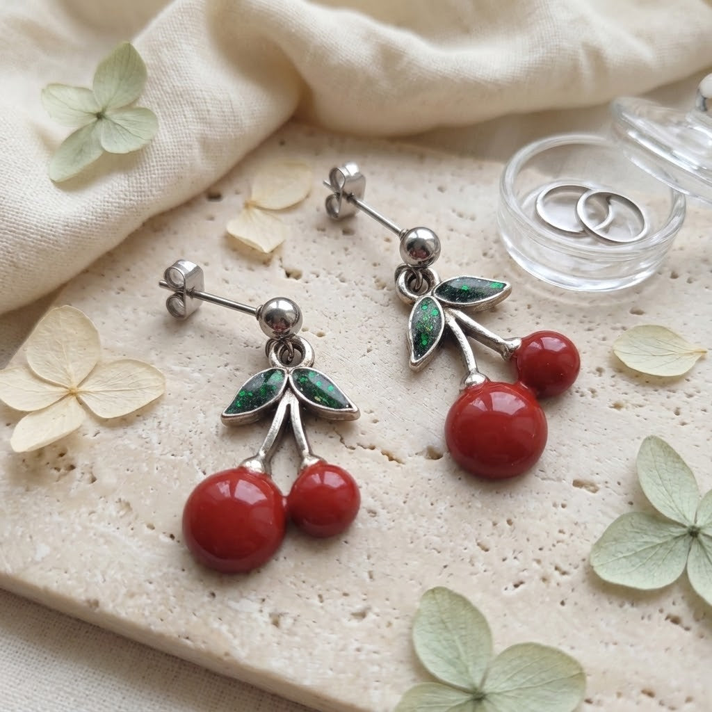 A pair of cherry drop earrings with hand-painted elements, green inlay leaves, and silver posts, displayed on a textured stone slab with dried flowers and a small glass ring box.