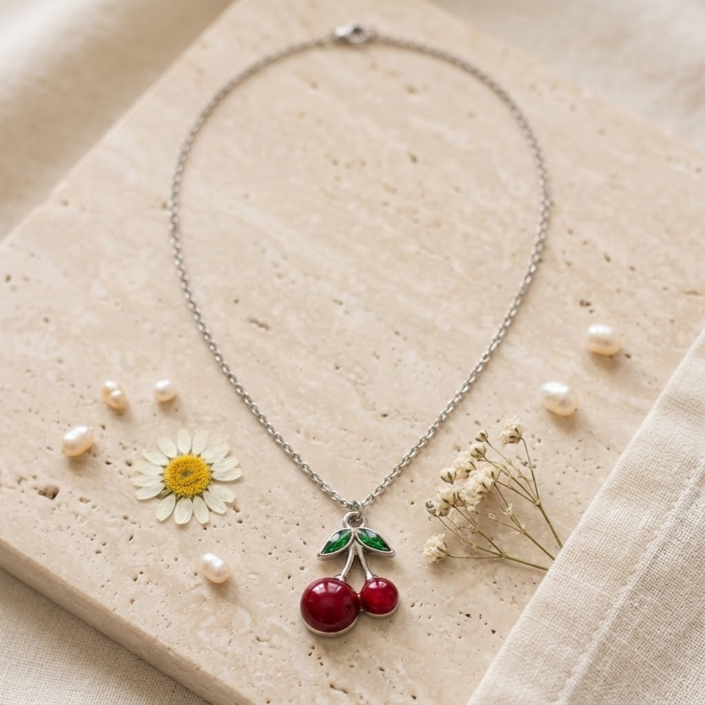 A silver-tone chain necklace with a cherry pendant, styled on a beige stone slab with small pearls, a daisy, and dried flowers.