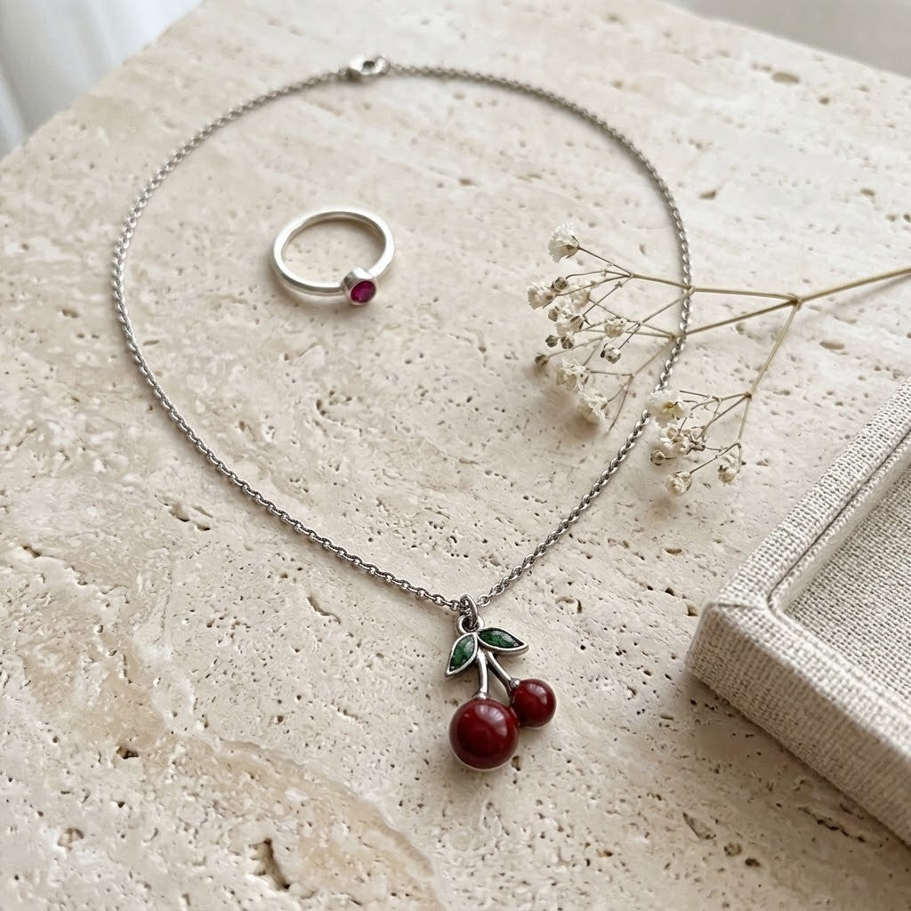 A silver-toned necklace with a cherry pendant and a matching silver ring, arranged on a textured beige stone surface with dried white flowers.