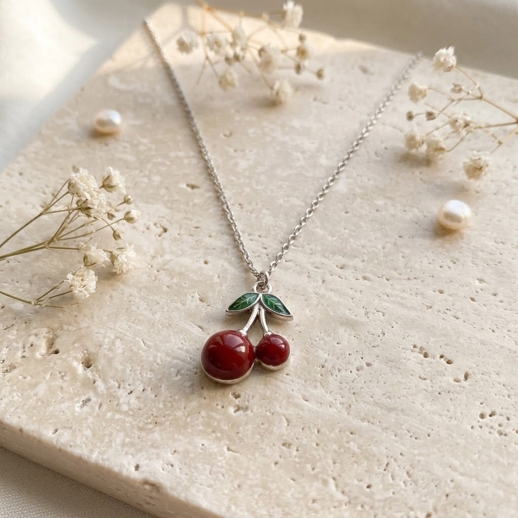 A silver necklace with a cherry pendant displayed on a textured beige stone slab with dried baby's breath and small pearls.