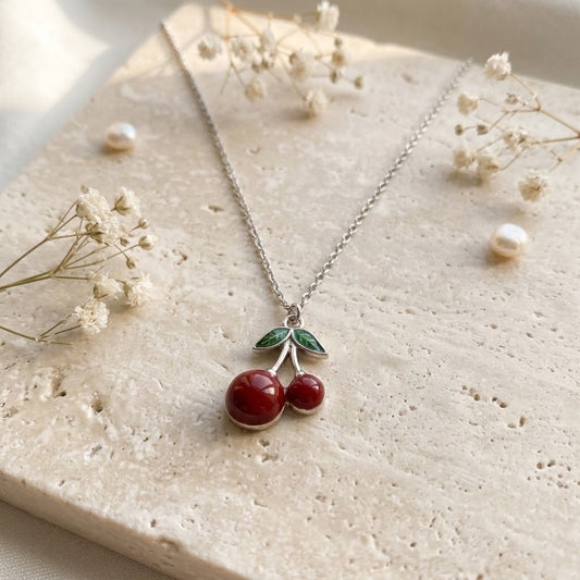 A silver necklace with a cherry pendant, featuring two red cabochon stones and green stone leaves, displayed on a textured beige stone slab with dried baby's breath and small pearls.