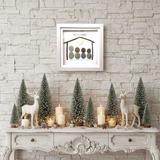 Family pebble art above decorative mantel with Christmas trees, candles, and deer figurines against a stone wall.