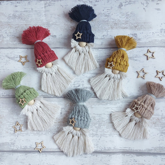 A collection of handmade Christmas gonk decorations made from high-quality single ply cotton twine, available in various colours, each adorned with a different embellishment, displayed on a white wooden background with stars and tassels.