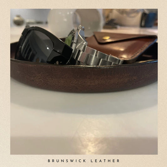 Brown leather tray with watches and a wallet on a reflective surface, branded 'Brunswick Leather'.