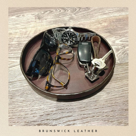 Round brown leather tray with sunglasses, watch, and keys on a wooden surface, branded 'Brunswick Leather'.