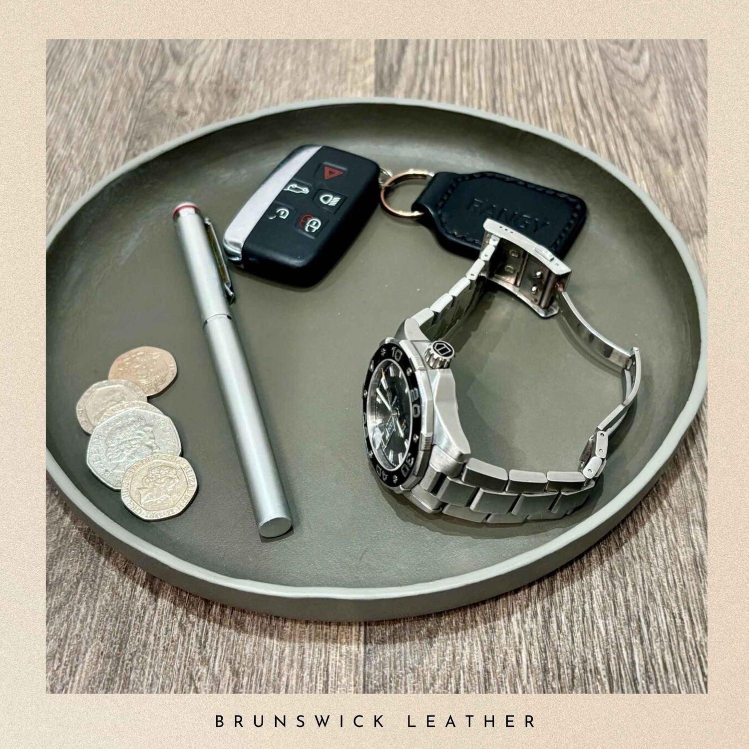 Silver watch, pen, car keys, and coins on a grey leather tray with 'Brunswick Leather' branding.