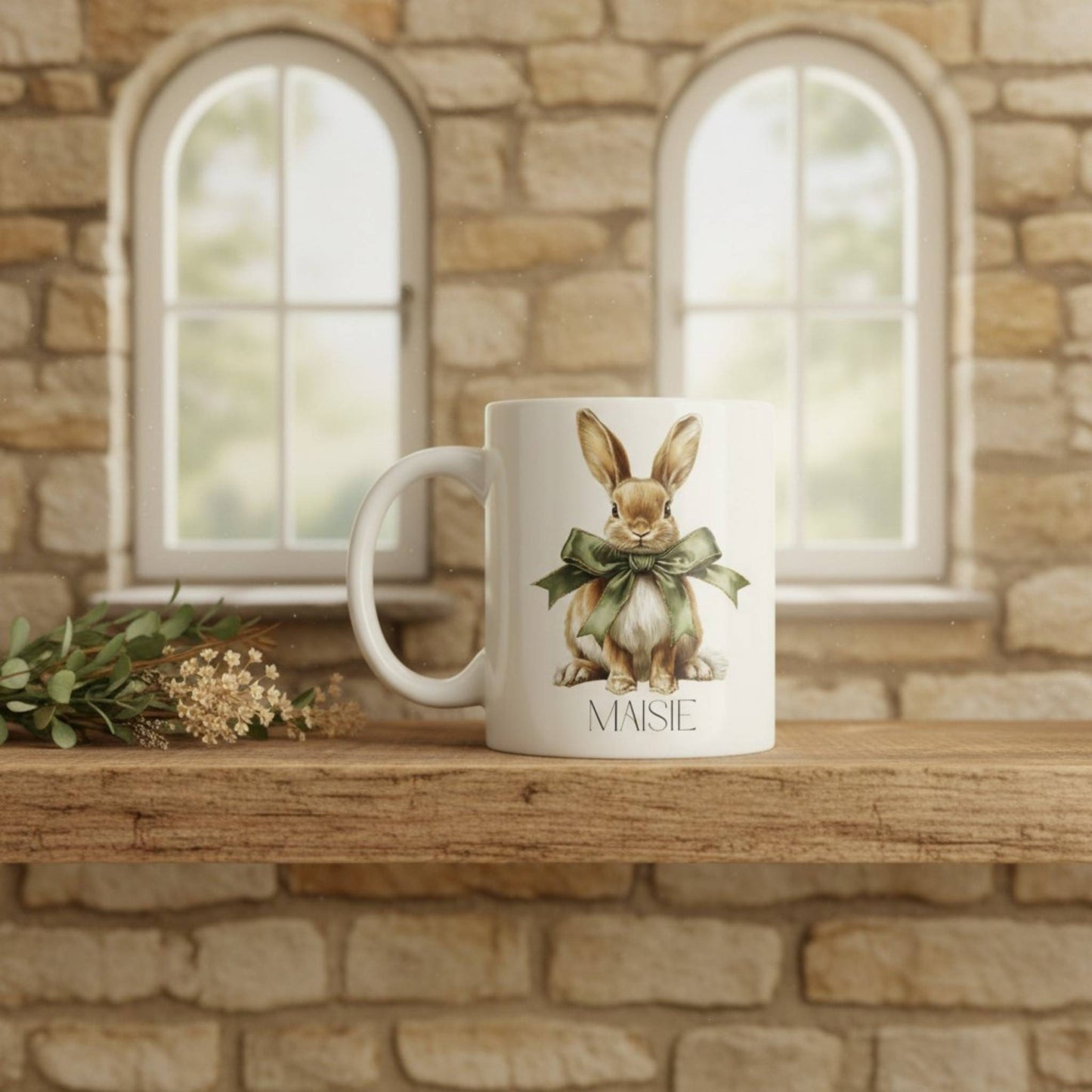 White mug with a hare design and 'Maisie' text on a wooden surface with stone wall background