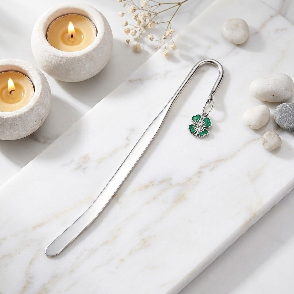 A silver metal bookmark with a green clover charm, styled on a white marble tray with lit candles, small stones, and dried flowers.