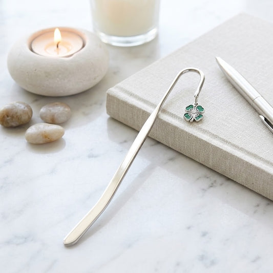 A silver metal bookmark with a small green clover charm rests on a beige linen notebook on a white marble surface, styled with a lit candle, stones, and a pen.