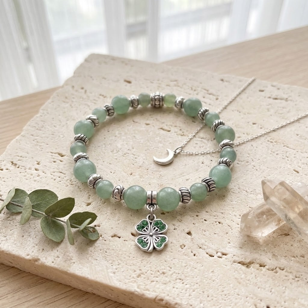 A beaded bracelet with light green jade-like beads, silver spacer beads, a silver crescent moon charm, and a silver four-leaf clover charm with green enamel details, displayed on a stone slab.