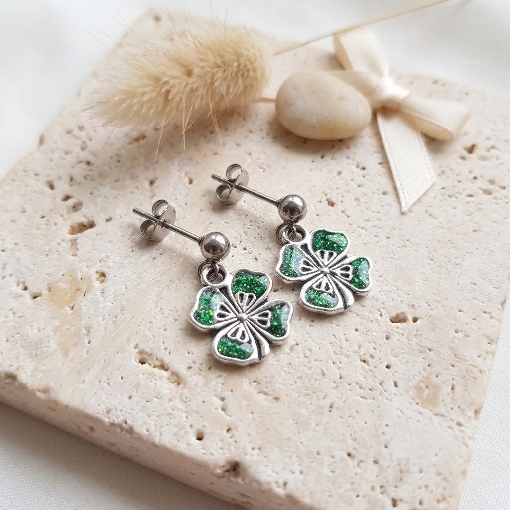 A pair of silver dangle earrings with green glittery clover charms, displayed on a textured beige stone slab with dried grass and a small ribbon.