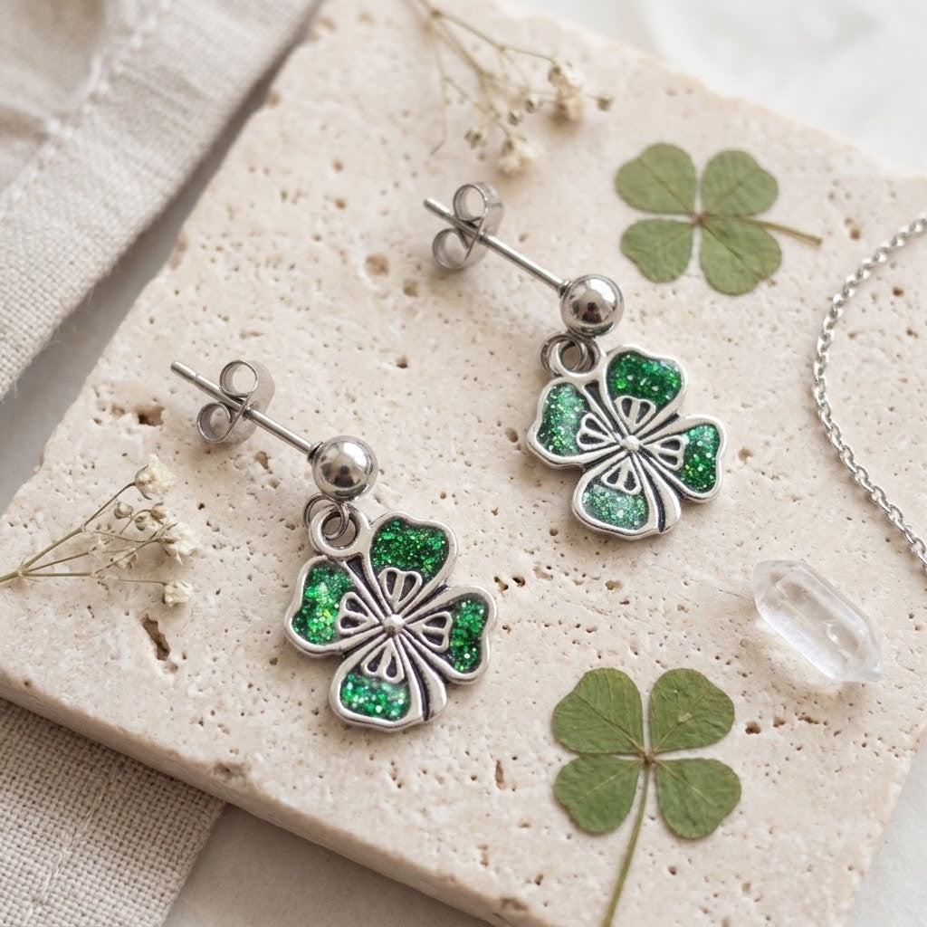 A pair of silver-toned dangle earrings with glittery green shamrock charms, displayed on a textured beige stone tile with dried flowers and green shamrock leaves.