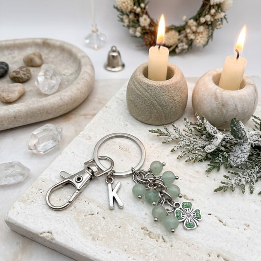 A silver-toned keychain with a lobster clasp, a letter K charm, light green bead charms, and a green clover charm, styled on a stone tray with candles, crystals, and greenery.