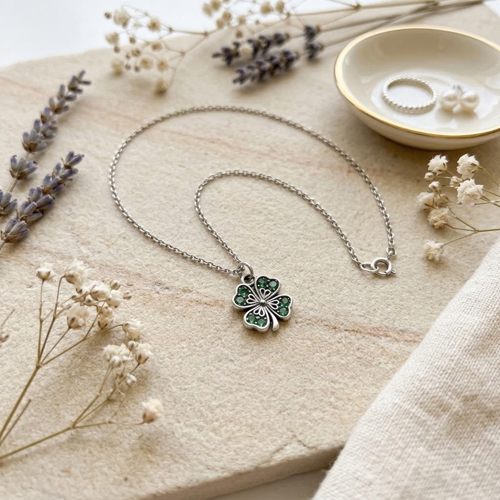 A silver chain necklace with a four-leaf clover pendant featuring green enamel and hand painted details, styled on a beige stone slab with dried flowers and a small dish of jewellery.