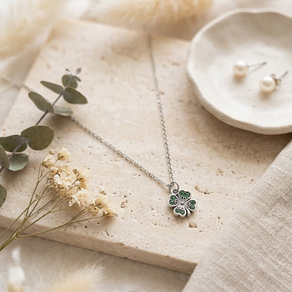 A delicate silver necklace with a green and silver four-leaf clover pendant, styled on a textured beige stone slab with dried flowers, eucalyptus leaves, and a small dish of pearl earrings.
