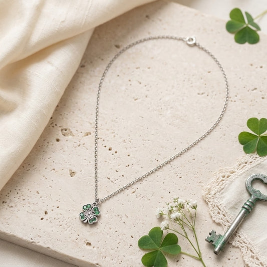 A silver chain necklace with a clover pendant featuring green enamel and pink crystal accents, styled on a textured beige stone slab with cream fabric, green clovers, and a vintage key.