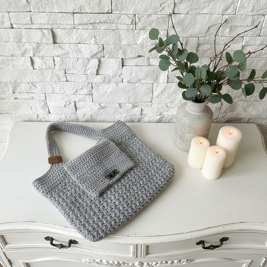 Grey textured handbag on a white surface with candles and a vase of greenery in the background.